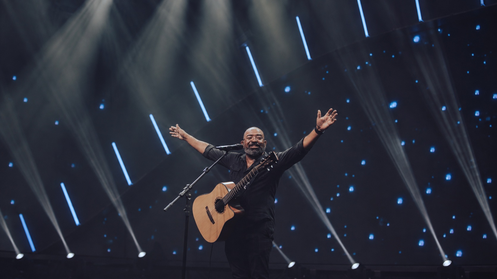 Luiz Arcanjo no palco, com violão e braços abertos sob luzes azuis